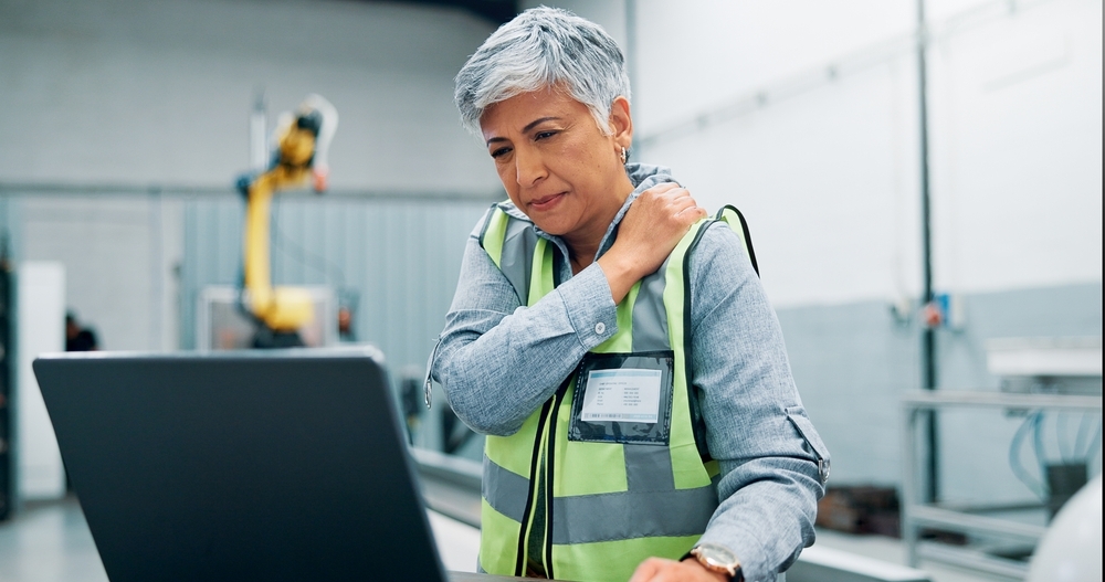 Female operator rubbing her shoulder in pain as she works on her laptop on the factory floor.